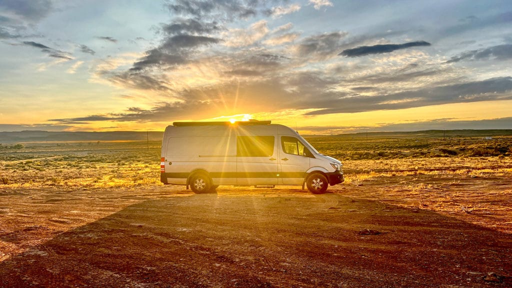 a silver Sprinter camper van is parked in an vast, open boondocking location in Moab, Utah with the sun setting behind the van. the sunset appears to be blasting out from the top of the van. Road trip apps.